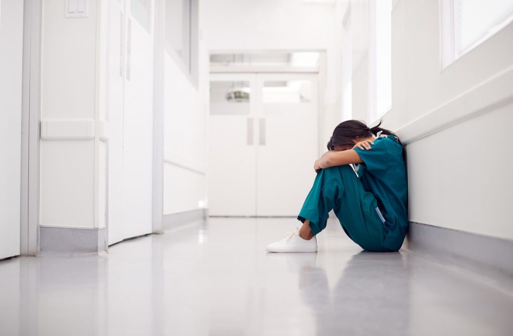 Shift Work Disorder Stressed And Overworked Nurse Wearing Scrubs Sitting On Floor In Hospital Corridor
