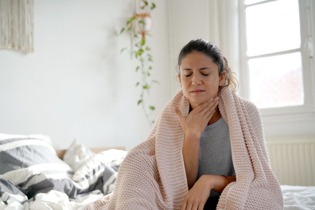 Waking Up With a Dry Throat: What Causes It and How Do You Treat It? A woman sits on bed and rubs her dry throat